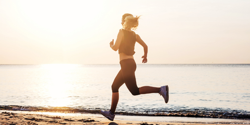 Keep Moving Woman wearing a black tank top and black leggings running of the beach at sunset.